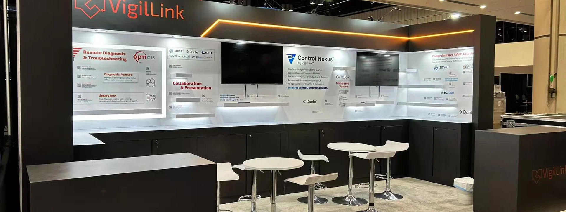 Completed VigilLink inline exhibit with dark illuminated walls, multiple screens, counters, and café tables.