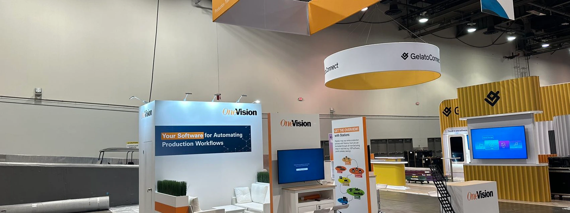 Completed OneVision island exhibit with a suspended sign, software demonstration screens, counters, and a small seating area.
