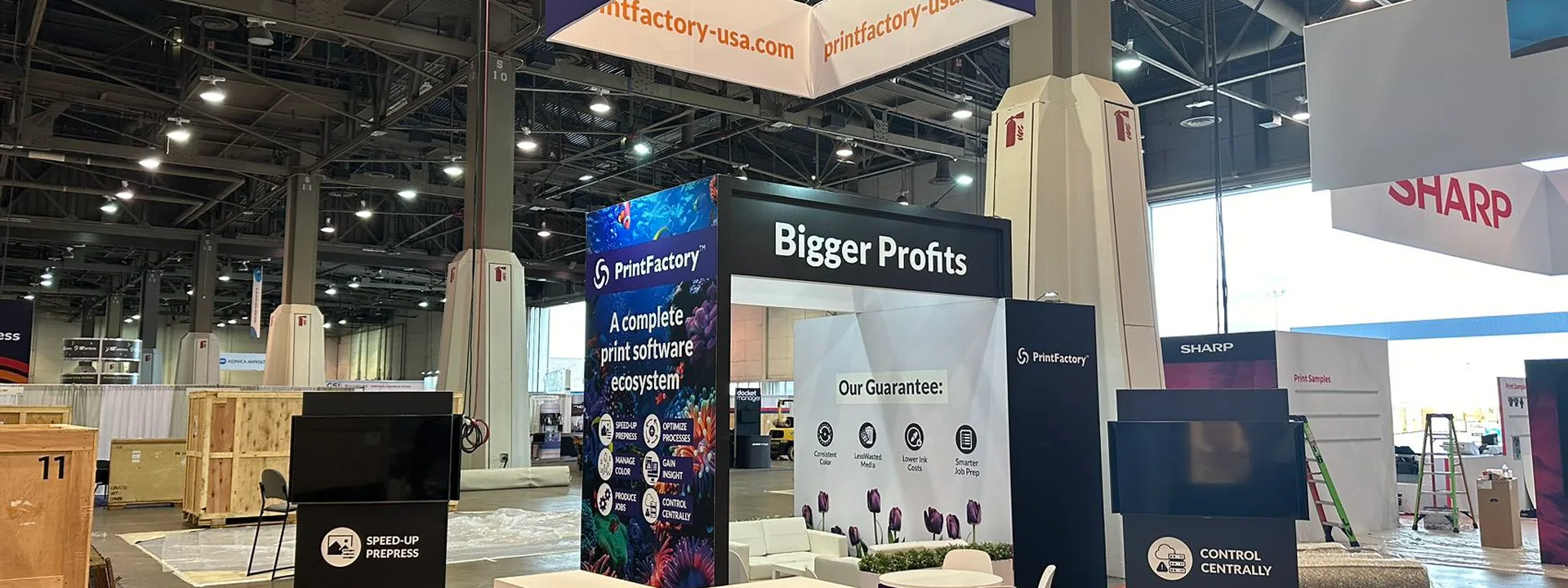 Completed PrintFactory island exhibit with an overhead sign, reception counter, display panels, and meeting chairs.