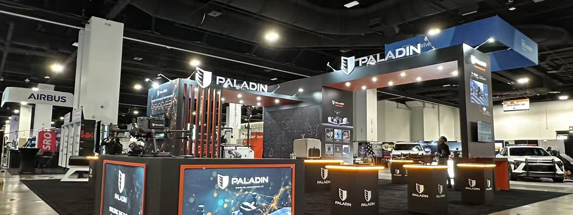 Large dark trade show exhibit with illuminated counters, overhead branding, product displays, and open aisles.